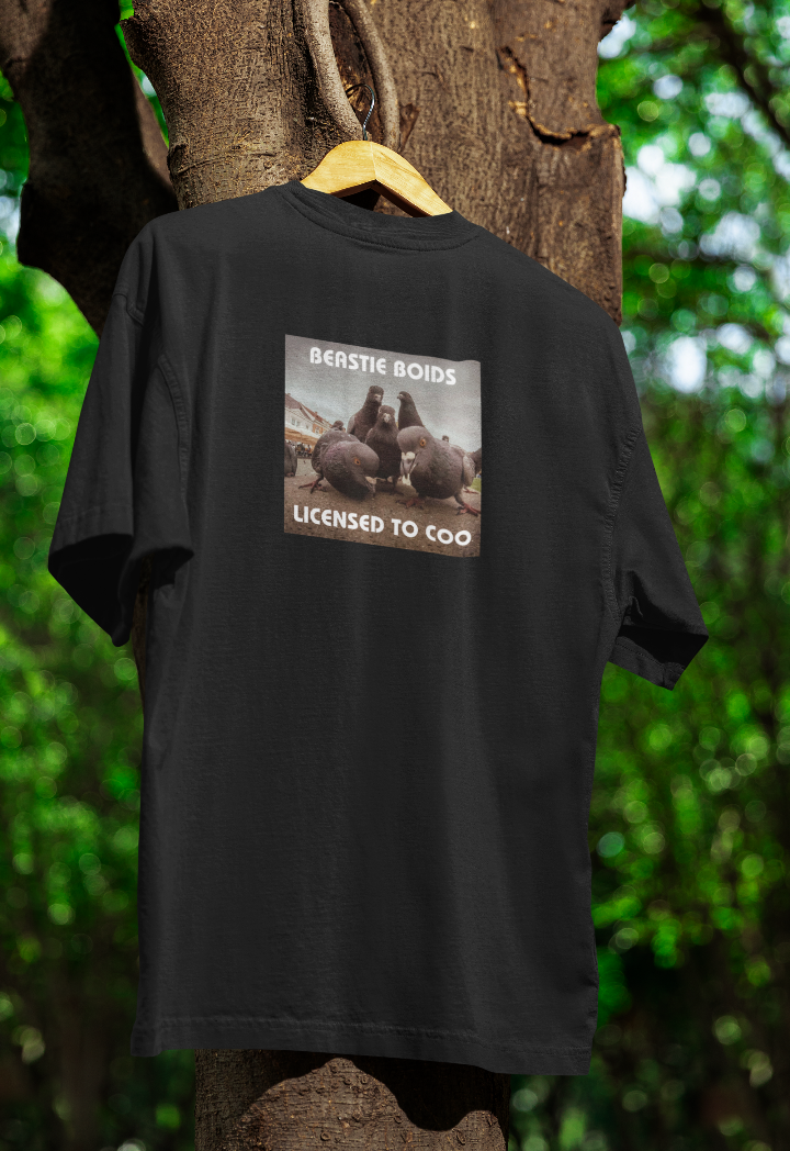 Black t-shirt hanging on a wooden hanger outdoors, featuring a parody album cover with pigeons labeled "Beastie Boids: Licensed to Coo." The image has a vintage, gritty aesthetic, showing pigeons in an urban setting with bold white text. The background features a tree with green foliage.