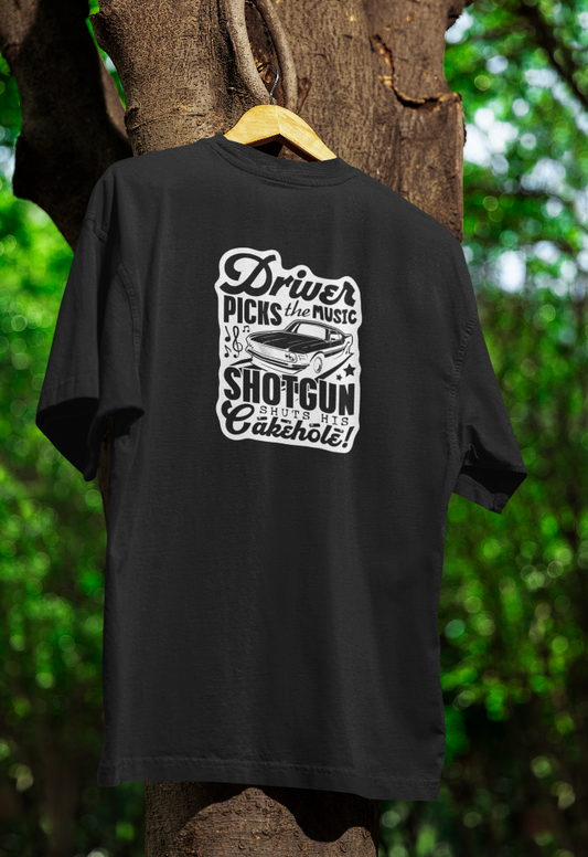 Black t-shirt hanging on a wooden hanger outdoors, featuring a white vintage-style design with the phrase "Driver picks the music, shotgun shuts his cakehole!" along with an illustration of a classic muscle car. The background includes a tree with green foliage.