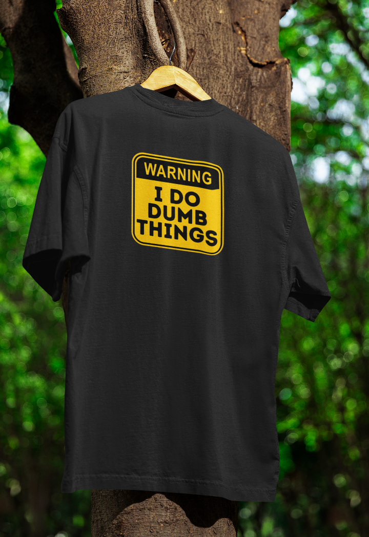 Black t-shirt hanging on a wooden hanger outdoors, featuring a bold yellow and black warning sign-style design with the text "WARNING: I DO DUMB THINGS." The background includes a tree with green foliage.