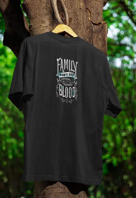 Black t-shirt hanging on a wooden hanger outdoors, featuring an ornate white and teal typography design that reads "Family Don’t End with Blood." The background includes a tree with green foliage.