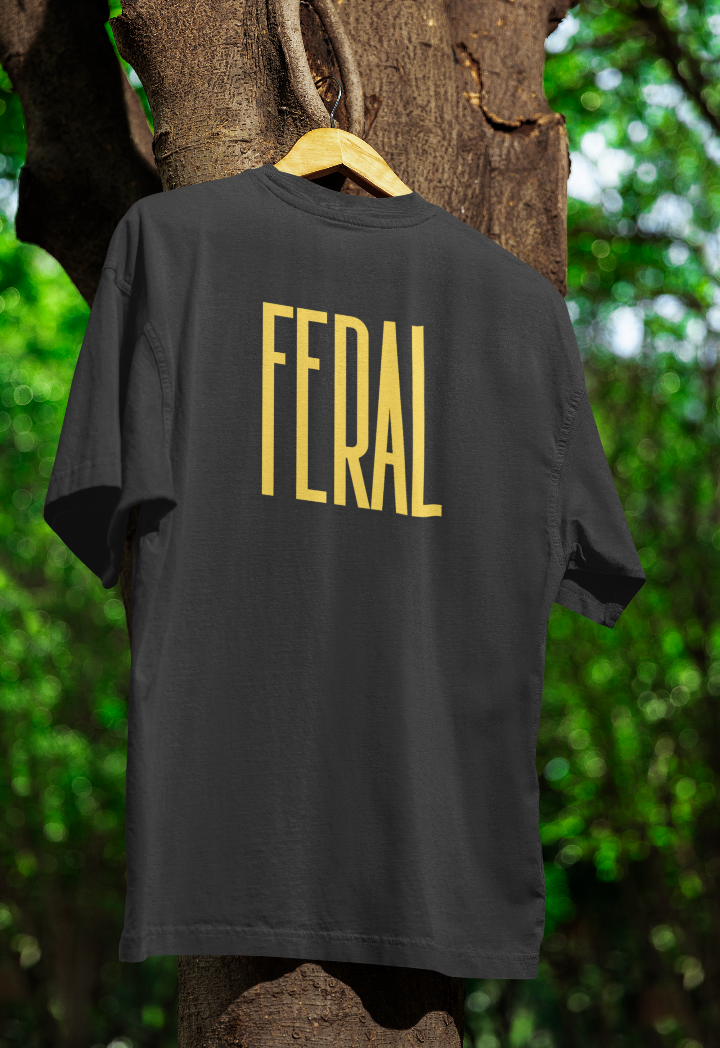 Black t-shirt with "FERAL" printed in bold yellow letters, hanging from a wooden hanger on a tree branch in an outdoor setting with lush green foliage in the background.