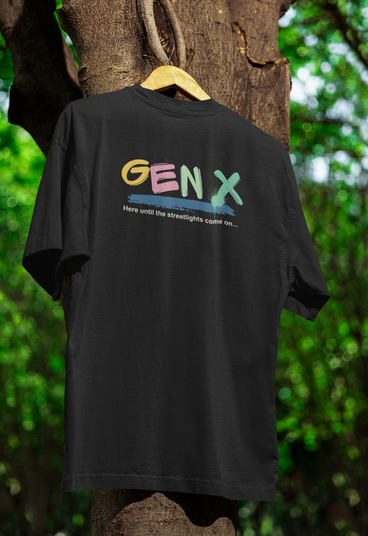 Black t-shirt hanging on a wooden hanger outdoors, featuring the text "GEN X Here until the streetlights come on..." in pastel and spray-paint-style lettering. The background includes a tree with green foliage.