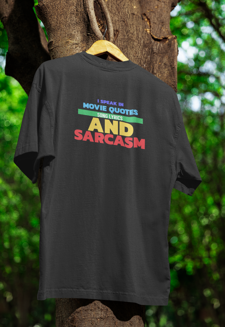 Black t-shirt hanging on a wooden hanger outdoors, featuring the text "I SPEAK IN MOVIE QUOTES, SONG LYRICS AND SARCASM" in bold, multicoloured block letters. The shirt is displayed against a tree with green foliage in the background.