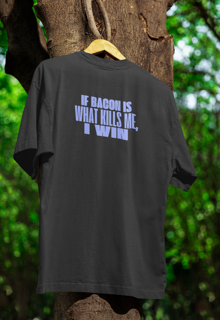 Black t-shirt hanging on a wooden hanger outdoors, featuring bold white text that reads "If bacon is what kills me, I win." The background includes a tree with green foliage.