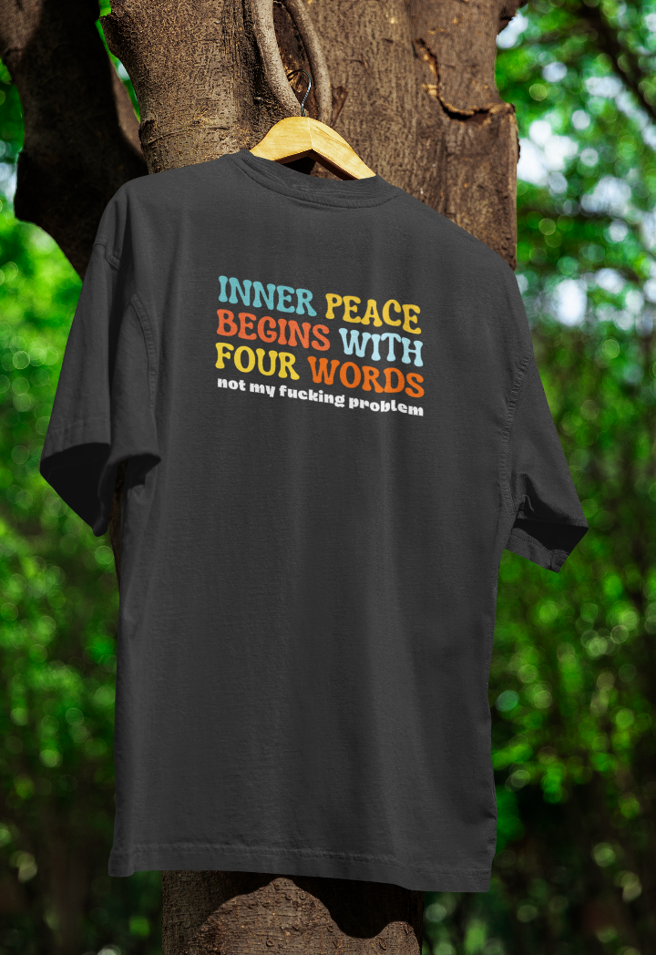 Black t-shirt hanging on a wooden hanger outdoors, featuring multicoloured text that reads "Inner peace begins with four words – not my f***ing problem." The background includes a tree with green foliage.