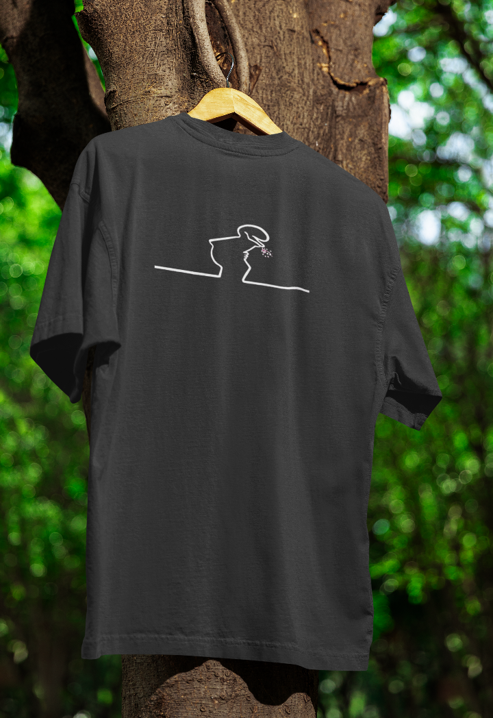 Black t-shirt hanging on a wooden hanger outdoors, featuring a minimalist white line drawing of a character with a tilted head, laughing with small dots representing a snorting effect. The background includes a tree with green foliage.