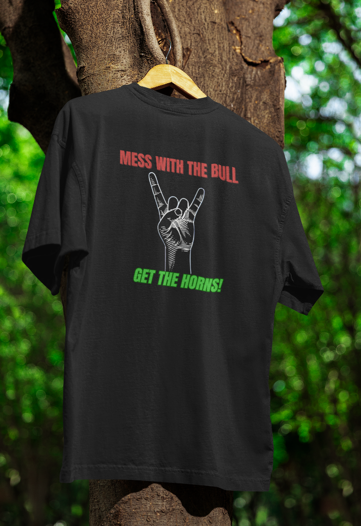 Black t-shirt hanging on a wooden hanger outdoors, featuring bold red and green text reading "Mess with the bull, get the horns!" with an illustration of a hand making the classic rock 'n' roll horns gesture. The background includes a tree with green foliage.