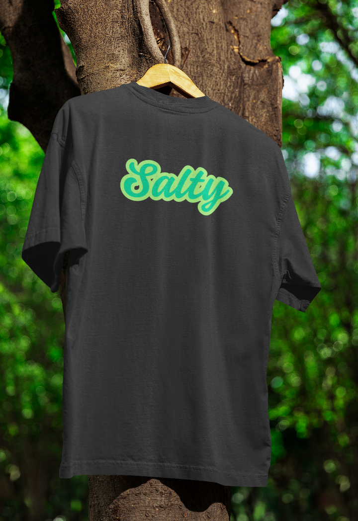 Black t-shirt hanging on a wooden hanger outdoors, featuring the word "Salty" in bold, playful green script. The background includes a tree with green foliage.