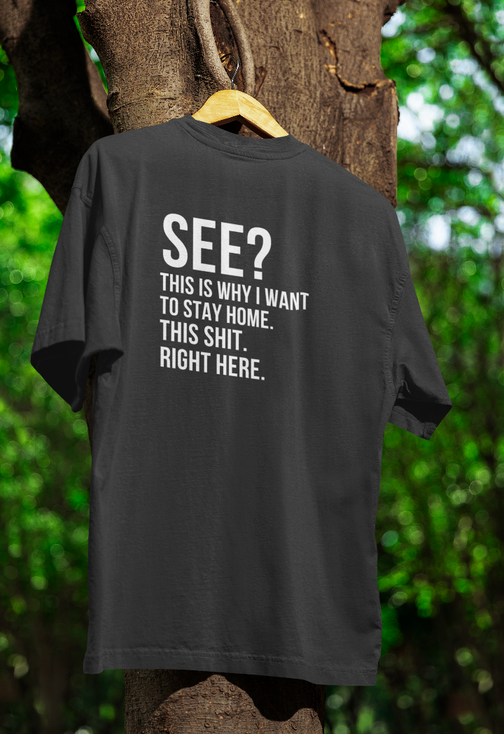 Black t-shirt hanging on a wooden hanger outdoors, featuring bold white text that reads: "SEE? This is why I want to stay home. This sh*t. Right here." The background includes a tree with green foliage.