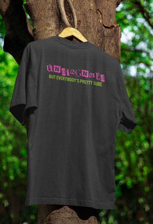 Black t-shirt hanging on a wooden hanger outdoors, featuring bold pink and green text that reads "Undiagnosed but everybody’s pretty sure," styled with a ransom-note-inspired font. The background includes a tree with green foliage.