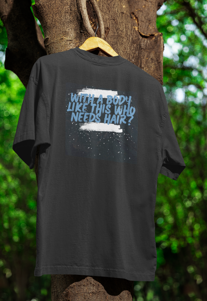 Black t-shirt hanging on a wooden hanger outdoors, featuring blue and white distressed-style text that reads, "With a body like this, who needs hair?" against a dark speckled background. The backdrop includes a tree with green foliage.